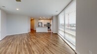 This single-floor unit is designed with a contemporary aesthetic, combining clean lines, neutral tones, and an abundance of natural light. The open-concept living and kitchen area is one of its most striking features, anchored by expansive floor-to-ceiling windows that wrap around a corner, offering panoramic urban views and flooding the space with daylight. A ceiling fan complements the airy atmosphere of the living area.

The kitchen is well-appointed with white cabinetry extending to the ceiling, stainless steel appliances including a French-door refrigerator, built-in microwave, and a gas range. A central island provides additional workspace and a natural division between the kitchen and living areas. Light wood-style flooring runs consistently throughout the main living spaces and bedrooms, adding warmth to the modern design.

The bedrooms are comfortably proportioned, each receiving natural light through standard windows with blinds. At least one bedroom includes a closet. The bathrooms reflect the same refined aesthetic — one features large-format white tiles, a frameless glass walk-in shower with a rainfall showerhead, and chrome fixtures, while the other offers a floating vanity with an illuminated mirror and light tile flooring, creating a clean, spa-like environment throughout.