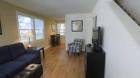 This two-floor residence offers a comfortable and functional layout designed with everyday living in mind. The main living area greets you with warm hardwood-style flooring that runs throughout, creating a cohesive and inviting atmosphere. Large windows along the exterior walls flood the space with abundant natural light, enhancing the bright, airy feel of the interior. The living room is thoughtfully arranged with seating and entertainment furnishings, including a media console and accent chair, while framed wall art adds a personalized touch to the neutral-toned walls. The living area flows naturally into a dedicated dining space, which benefits from its own set of windows overlooking the outdoors, making it an ideal spot for meals throughout the day. A dining table with coordinating chairs anchors this area, reinforcing the open and connected feel between the two spaces. The warm yellow accent walls in the dining area contrast pleasantly with the crisp white tones of the living room, adding visual interest and depth. Spanning two floors, the residence provides ample space for both relaxing and entertaining, with a layout that balances openness with defined functional zones. The overall ambiance is welcoming, bright, and well-maintained.