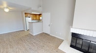 This single-floor residence offers a thoughtfully designed open layout that seamlessly connects the living and kitchen areas. The living room features soaring vaulted ceilings that lend an airy, spacious feel to the space, complemented by light wood-look flooring that extends throughout. A white-tiled fireplace with a black insert serves as a distinctive focal point along one wall, adding warmth and character to the room.

The kitchen is accessed via a raised pass-through counter finished with a granite-style surface, providing both a functional workspace and a casual dining or serving area. Warm honey-toned wood cabinetry lines the kitchen walls, offering ample storage, while a white refrigerator and under-cabinet lighting contribute to a clean, practical aesthetic. A pendant light fixture above the pass-through adds a finishing touch to the kitchen zone.

Neutral beige walls and white trim run throughout, creating a bright and cohesive backdrop that suits a variety of décor styles. A white paneled entry door opens directly into the main living space, enhancing accessibility and flow. The overall ambiance is clean, open, and move-in ready, making this a versatile and welcoming single-level home.