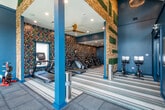 This single-floor fitness center is thoughtfully designed to accommodate a full range of workout needs within a visually striking environment. The space is divided into two primary zones: a dedicated cardio area and a strength training floor, each with distinct flooring to suit their purpose. The cardio zone features a row of modern treadmills and stationary bikes arranged along a bold, multicolored patterned accent wall, complemented by striped carpet flooring and recessed lighting. Large ceiling fans contribute to a comfortable workout environment throughout. The strength training area transitions to durable rubber flooring and benefits from soaring vaulted ceilings with large floor-to-ceiling windows that flood the space with natural light and offer views of the surrounding greenery. This zone is equipped with a red power rack, a functional cable machine, a dumbbell rack, and additional free weights. A wall-mounted flat-screen TV adds a multimedia element to the workout experience. The entryway makes a strong first impression with its vibrant wallpaper, warm wood flooring, a sleek dark credenza, a gold-framed circular mirror, and floral accents—setting an elevated, design-forward tone that carries throughout the entire facility.