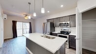 This contemporary single-floor residence showcases a thoughtfully designed open-concept layout that seamlessly connects the kitchen and living areas. The kitchen is a standout feature, centered around a large island with a quartz countertop, double undermount sink, and sleek gray cabinetry. A full suite of stainless steel appliances — including a refrigerator, electric range, and built-in microwave — adds a polished, modern touch, while pendant lighting above the island and recessed ceiling lights create a well-lit, inviting atmosphere. A walk-in pantry provides generous storage space. Wood-look plank flooring flows throughout the main living areas, complemented by a ceiling fan that enhances comfort. The living area opens directly to a private balcony through sliding glass doors, filling the space with natural light. The bedrooms are finished with plush carpet and individual wall-mounted mini-split HVAC units for personalized climate control, with windows offering exterior views. The bathrooms feature dark wood vanities with white countertops, framed mirrors, and wood-look tile flooring, maintaining the home's cohesive, modern aesthetic. Clean white walls and crisp trim throughout contribute to a bright, fresh ambiance.