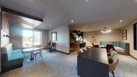 This thoughtfully designed single-floor communal space blends contemporary aesthetics with functional versatility. The open-plan layout is anchored by a large dark conference-style table with tan leather chairs at its center, ideal for group work or meetings. Adjacent to this is a casual lounge corner featuring a teal upholstered booth and accent chairs in red and neutral tones, framed by colorful artwork that adds a vibrant focal point to the warm, neutral-toned walls.

A dedicated beverage and refreshment station, set against a striking geometric tile accent wall in dark tones, provides a practical hub for gatherings. Nearby, high-top bar seating positioned along a window wall allows natural light to pour into the space, offering views of the surrounding outdoor greenery. A decorative ceiling element with metallic geometric tiles adds an architectural flourish overhead.

The flooring throughout features a textured, speckled carpet that contributes to the space's comfortable, modern character. Glass-paneled partition walls and full-length glass entry doors line the corridor, maintaining an open, transparent feel while defining separate zones. Recessed lighting throughout ensures a well-lit, inviting ambiance. The overall design balances professional functionality with a welcoming, social atmosphere.