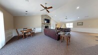 Located at 885 Castlewood Blvd in Charleston, South Carolina, this single-story community clubhouse offers a warm and welcoming gathering space designed for comfort and socializing. The main room is defined by soaring vaulted ceilings that lend an open, airy feel, complemented by ceiling fans that keep the space comfortable year-round. A striking floor-to-ceiling stacked stone fireplace serves as the room's dramatic focal point, topped with a mounted flat-screen television and a rustic wood mantel, making it both a functional and aesthetic centerpiece. Plush, patterned carpeting in warm neutral tones runs throughout, adding to the inviting atmosphere. The seating area features generous leather sofas arranged around a classic wooden coffee table, providing ample space for gatherings. Adjacent to the lounge area is a dining section with a wooden table and chairs, ideal for casual meals or meetings. A kitchenette area with cabinetry offers convenient amenities for events and gatherings. White wainscoting lines the walls, adding a refined architectural detail, while recessed lighting and natural light from multiple windows brighten the interior. A bulletin board and desk area provide a practical resource corner, rounding out this well-appointed community space.
