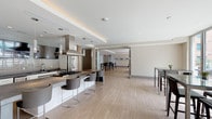 Located in Hughes Landing, Spring, TX, this single-floor amenity space is designed with a contemporary aesthetic and offers a well-appointed environment for gathering and entertaining. The open-plan layout is anchored by a sleek, fully equipped communal kitchen featuring stainless steel appliances, a large French-door refrigerator, dual professional-grade range hoods, a subway tile backsplash, and a substantial island with a concrete-style countertop and bar seating for multiple guests. Pendant lighting and cove lighting accent the coffered ceiling above, adding warmth to the otherwise clean, modern palette of whites, grays, and light wood-tone flooring that extends throughout the space. Adjacent to the kitchen, a dining area provides high-top tables and seating for casual meals or social gatherings. Framed abstract artwork adds a curated decorative touch along the neutral-toned walls. Glass-panel sliding doors lead seamlessly to an outdoor patio, where high-top seating continues al fresco. Floor-to-ceiling windows flood the space with natural light and frame views of a resort-style pool, palm trees, and the surrounding residential community. The overall ambiance is polished, functional, and inviting.