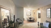 This single-floor residence offers a thoughtfully designed open-concept layout that connects the living and dining areas in a seamless, functional flow. The living area features plush carpeting underfoot, providing a warm and comfortable atmosphere, while neutral gray and beige tones across the walls create a calm, cohesive aesthetic throughout the space. A decorative accent wall in the dining area adds a touch of visual interest, complementing the ceiling fan overhead that serves both as a functional fixture and a subtle design element. The dining space comfortably accommodates a round table with seating for multiple guests, making it well-suited for both everyday meals and casual entertaining. Textured ceilings add depth and character to the interior, while the open passageways between rooms contribute to an airy, connected feel. Tasteful decorative accents — including framed wall art, a sculptural wall piece, and floral arrangements — are thoughtfully placed throughout, enhancing the overall warmth of the space. Hardwood-style flooring is visible in the dining area, offering a pleasing contrast to the carpeted living space. The overall ambiance is welcoming, practical, and move-in ready.