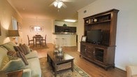 This single-floor residence offers an open-concept layout that seamlessly connects the living, dining, and kitchen areas, creating a cohesive and functional living environment. The living area is anchored by warm hardwood flooring and features a ceiling fan, crown molding, and ample space for comfortable furnishings. A large entertainment center with rich, dark wood cabinetry serves as a focal point of the room, complemented by a patterned area rug that adds texture and warmth to the space. Adjacent to the living area, the dining space is illuminated by an elegant chandelier and provides a welcoming setting for meals and gatherings. The kitchen is thoughtfully designed with dark cabinetry, modern appliances including a built-in microwave, and a raised breakfast bar with bar stool seating — ideal for casual dining. Crown molding throughout adds a refined architectural touch, while the warm, neutral wall tones create a cohesive and inviting ambiance. The open flow between all areas enhances the sense of space, making this residence both practical and comfortable for everyday living.