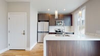 This single-floor space showcases a cohesive modern design throughout, combining clean lines, quality finishes, and an open, functional layout. The kitchen serves as a central highlight, featuring rich dark wood cabinetry that extends to the ceiling, providing generous storage. A large island with a crisp white quartz waterfall countertop anchors the space, equipped with an undermount sink and a sleek industrial-style faucet. Stainless steel appliances, including a French door refrigerator, electric range, and built-in microwave, complement the contemporary aesthetic. Cylindrical glass pendant lights hang above the island, adding a refined touch. Light natural hardwood flooring runs consistently throughout the unit, enhancing the sense of warmth and continuity. Additional rooms offer bright, open spaces filled with natural light from large windows fitted with horizontal blinds. The bathroom is notably luxurious, featuring floor-to-ceiling marble-look porcelain tile, a frameless glass walk-in shower with a multi-function chrome fixture system, a wood-grain vanity, and marble-topped counters. Recessed lighting and neutral wall tones throughout create a clean, move-in-ready atmosphere.