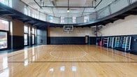 Located at 133 S Ashland Ave in Chicago's West Loop neighborhood, this impressive indoor basketball facility showcases a thoughtfully preserved and modernized athletic space. The centerpiece is a full regulation hardwood basketball court, finished to a high gloss and marked with crisp court lines, anchored by a bold custom logo at center court featuring a black and gold referee mascot design.

The soaring ceiling height accommodates regulation play with ease, and the space is further defined by original exposed brick walls finished in a two-tone treatment — natural brick above and painted black wainscoting below — lending a classic gymnasium character. A curved wraparound mezzanine level, framed by black steel railings and safety netting, overlooks the court and is accessible via a dramatic spiral staircase in the corner. Vintage "HOME" and "VISITORS" scoreboards add an authentic competitive atmosphere.

Natural light pours in through large windows along one side of the court, which also provide views to an adjacent outdoor terrace. Overhead, industrial-style pendant lighting supplements the natural light throughout the space. A dumbbell and equipment rack along the sideline rounds out the athletic amenities. The combination of historic architectural elements with modern court finishes makes this a standout facility for sport and recreation in Chicago.