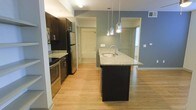This single-floor residence showcases a thoughtfully designed open-concept layout that seamlessly connects the kitchen, dining, and living areas. The kitchen is a standout feature, outfitted with rich dark cabinetry that contrasts elegantly against light granite countertops. A central island with an integrated sink serves as both a functional workspace and a social hub, illuminated by a trio of sleek pendant lights that add a contemporary touch. Stainless steel appliances, including a refrigerator, stove, and microwave, complete the well-appointed culinary space.

Light hardwood flooring flows throughout, lending warmth and continuity to the open floor plan. The walls are finished in a muted slate blue accent, adding depth and a modern aesthetic to the space. Natural light filters in through windows visible in the adjacent rooms, enhancing the bright and airy atmosphere.

Beyond the kitchen, the layout opens toward additional rooms, offering a glimpse of well-proportioned living and bedroom spaces. The clean architectural lines, neutral palette, and quality finishes throughout reflect a cohesive and contemporary design sensibility, making this a functional and visually appealing residence.