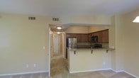 This single-floor residence offers a well-designed open layout that seamlessly connects the living area and kitchen. The kitchen is a standout feature, equipped with stainless steel appliances — including a full-size refrigerator and microwave — warm-toned wooden cabinetry, and granite countertops that extend to a raised breakfast bar with shelved support, ideal for casual dining or additional workspace. The flooring transitions between tile in the kitchen and entry areas to carpet throughout the living and hallway spaces, providing both practicality and comfort. A central hallway extends from the main living area, lined with doors that lead to additional rooms, suggesting a thoughtful layout that separates living and private spaces. Ceiling-mounted light fixtures provide consistent illumination throughout, while HVAC vents are integrated into the ceiling, ensuring climate control across the space. The neutral, warm beige tones on the walls and trim create a cohesive and inviting ambiance throughout the entire residence. The overall design balances functionality with a welcoming aesthetic, making it a comfortable and practical living environment.