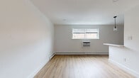 This single-floor space offers a bright and clean aesthetic throughout, characterized by crisp white walls, smooth ceilings with recessed lighting, and warm light wood laminate flooring that runs continuously across the main living areas. The open living area is well-proportioned, bathed in natural light from a window fitted with white blinds, and is equipped with a wall-mounted air conditioning unit and baseboard heating for year-round comfort. Two interior doors lead to additional spaces, providing a functional layout. The kitchen is a standout feature, showcasing white shaker-style cabinetry with matte black hardware, a light stone-toned countertop, and a herringbone-patterned light blue tile backsplash. It is outfitted with stainless steel appliances, including a gas range and over-the-range microwave, along with a matte black pull-down faucet over an undermount sink. The bathroom is thoughtfully designed with dark slate-toned floor tiles, a full bathtub and shower combination, and a bold geometric patterned wall tile in muted blue-green tones, complemented by dark fixtures that add a modern edge. Overall, the space presents a cohesive, updated interior with a neutral palette and contemporary finishes throughout.