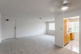 This single-floor residence offers a practical and comfortable layout designed for everyday living. The open living area is generously proportioned, finished with neutral-toned carpeting and cream-colored walls that create a bright, versatile backdrop. A ceiling fan with a light fixture provides overhead illumination and air circulation, while a front entry door opens directly into this main living space. A window along the back wall allows natural light to filter in.

Adjacent to the living area, the galley-style kitchen is efficiently arranged with warm honey-toned wood cabinetry lining both walls, offering ample upper and lower storage. Dark-veined countertops provide workspace, and white appliances including a refrigerator and dishwasher are included. Light tile flooring runs throughout the kitchen, transitioning to carpet in the adjoining dining or flex area nearby.

The bedrooms are carpeted and well-sized, each featuring windows that bring in outdoor views and natural light. Ceiling fans or flush-mount light fixtures are present in the sleeping areas. The bathroom includes a full tub-shower combination with white tile surround, a toilet, and a vanity with a single sink and mirror, all finished in a clean, neutral palette throughout.