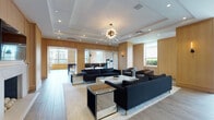 Located at 65 Bay St in Jersey City, NJ, this single-floor amenity space offers a refined and contemporary environment defined by warm wood-paneled walls, light hardwood-style flooring, and a thoughtfully curated interior aesthetic. The expansive main lounge area is anchored by a striking starburst chandelier suspended from a coffered ceiling, casting an elegant glow over a generous seating arrangement of navy blue sofas, leather armchairs, and mirrored accent tables. Recessed lighting throughout enhances the bright, airy atmosphere. Large windows allow abundant natural light to fill the space, framing views of the surrounding Jersey City streetscape and lush greenery. A secondary seating alcove near glass entry doors provides a welcoming transition area, furnished with plush velvet chairs and contemporary side tables. The fully equipped kitchen features high-gloss slate-blue cabinetry, a deep blue subway tile backsplash, premium stainless steel appliances including a Viking range and French door refrigerator, and an adjoining dining area with expansive floor-to-ceiling windows that capture sweeping views of the Hudson River and surrounding skyline. The overall ambiance is polished and functional, ideal for gathering and entertaining.