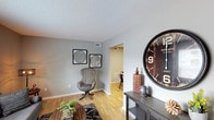 This single-floor, 2-bedroom, 1.5-bathroom residence offers a thoughtfully arranged layout that maximizes functionality without sacrificing comfort. The living area sets a welcoming tone with warm hardwood flooring, soft gray walls, and a textured ceiling that adds subtle depth overhead. A charming accent wall display, decorative furnishings, and an oversized statement clock contribute to the space's curated, lived-in character. Visible HVAC vents throughout indicate a central air system for year-round comfort. An open passageway connects the living room to a dining area, creating a natural flow between the main gathering spaces. White trim and baseboards provide clean contrast against the neutral gray walls, lending the interior a crisp, polished finish. The half bathroom adds everyday convenience for guests, while the two bedrooms offer private retreats. The overall ambiance is casual yet stylish, blending practical design with warm, inviting details. This residence is well-suited for those seeking a compact, move-in ready home with a coherent aesthetic and efficient use of space throughout its single level.