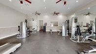 Located at 4500 Baymeadows Rd in Jacksonville, Florida, this fitness center offers a well-appointed exercise space designed to support a well-rounded workout routine. The main exercise room is generously sized, featuring durable speckled flooring that is both practical and easy to maintain. The space is outfitted with a comprehensive selection of equipment, including treadmills, elliptical machines, cable strength machines, and a dumbbell rack with a flat weight bench, providing options for both cardiovascular and resistance training. Recessed lighting and multiple ceiling fans with red-accented blades keep the environment well-lit and comfortable. A large wall mirror enhances the sense of openness, while a mounted flat-screen TV adds convenience for extended workout sessions. French doors along one wall allow natural light to filter in and provide a view of the surrounding outdoor area. A pass-through window connects to an adjacent room, offering added functionality. The secondary room features a carpeted floor and serves as a flexible multipurpose space. Together, the two areas create a complete fitness facility that is clean, organized, and ready to accommodate a variety of exercise needs.