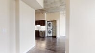 This well-appointed two-bedroom, one-bathroom unit offers a generous single-floor layout defined by contemporary industrial-modern design. Exposed concrete ceilings throughout lend an urban aesthetic, complementing the dark hardwood-style flooring found across the main living areas and kitchen.

The kitchen is outfitted with dark espresso cabinetry, white quartz countertops, a textured tile backsplash, and a full suite of stainless steel appliances including a refrigerator, range, built-in microwave, and dishwasher. A stacked washer and dryer unit is conveniently integrated near the kitchen area within a dedicated closet.

The open living area features floor-to-ceiling windows and sliding glass doors that lead to a balcony, offering expansive city skyline views and abundant natural light. The generously sized primary bedroom continues the concrete ceiling aesthetic, with carpet flooring, balcony access, a ceiling fan, and a large walk-in closet. The second bedroom also offers carpet flooring and city views. Both closets feature wrap-around shelving and hanging rods.

The bathroom includes wood-toned cabinetry, a white countertop vanity, a framed mirror, and hardwood-style flooring, presenting a clean, modern finish throughout.