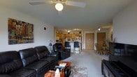 This single-floor residence offers an open-concept layout that seamlessly connects the living, dining, and kitchen areas into one cohesive and functional space. The living area is anchored by plush leather reclining sofas arranged around a decorative area rug, with a ceiling fan overhead providing both lighting and airflow. A coffee table and tasteful wall art add warmth and character to the space.

The kitchen is positioned toward the rear of the open floor plan and features light wood cabinetry, a white refrigerator, and a built-in microwave. Track lighting illuminates the workspace, and a raised breakfast bar with bar stools provides a convenient spot for casual dining or entertaining. Adjacent to the kitchen, a dedicated dining area accommodates a wooden table and chairs, offering a more formal setting for meals.

The flooring throughout is a neutral-toned carpet that adds comfort underfoot and ties the various living zones together. The interior doors are finished in warm wood tones, complementing the kitchen cabinetry. The overall palette is light and neutral, creating a bright, welcoming ambiance. The layout is efficiently designed to maximize usable space while maintaining a comfortable and inviting atmosphere suitable for everyday living.