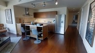 This single-floor space showcases a thoughtfully designed open-concept kitchen that combines contemporary style with functional living. The kitchen is anchored by a substantial central island finished in warm walnut-toned wood, which doubles as a casual dining area with modern swivel bar stools in a brushed metal finish. Rich, dark hardwood flooring extends throughout, adding warmth and visual continuity to the space.

The cabinetry features a consistent dark wood finish across both upper and lower units, providing ample storage while maintaining a clean, cohesive aesthetic. A stainless steel range hood is prominently positioned above the cooktop, complementing the full-size stainless steel French door refrigerator with built-in water and ice dispenser. Under-cabinet lighting brightens the workspace, while adjustable track lighting mounted on the ceiling allows for flexible illumination throughout.

The open layout creates a natural flow between the kitchen and the surrounding living areas, with the hardwood floors and neutral wall tones contributing to an overall atmosphere that is both welcoming and refined. The design balances practicality with modern elegance.