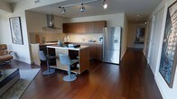 This single-floor space showcases a thoughtfully designed open-concept kitchen that combines contemporary style with functional living. The kitchen is anchored by a substantial central island finished in warm walnut-toned wood, which doubles as a casual dining area with modern swivel bar stools in a brushed metal finish. Rich, dark hardwood flooring extends throughout, adding warmth and visual continuity to the space.

The cabinetry features a consistent dark wood finish across both upper and lower units, providing ample storage while maintaining a clean, cohesive aesthetic. A stainless steel range hood is prominently positioned above the cooktop, complementing the full-size stainless steel French door refrigerator with built-in water and ice dispenser. Under-cabinet lighting brightens the workspace, while adjustable track lighting mounted on the ceiling allows for flexible illumination throughout.

The open layout creates a natural flow between the kitchen and the surrounding living areas, with the hardwood floors and neutral wall tones contributing to an overall atmosphere that is both welcoming and refined. The design balances practicality with modern elegance.