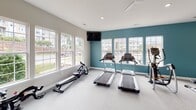 Located at 180 Embassy Dr in Fort Mill, SC, this single-floor fitness center is thoughtfully designed to support a wide range of workout routines. The space is divided into distinct functional zones, each tailored to a specific type of exercise. The cardio area is lined with large windows that flood the room with natural light and offer views of the surrounding community, creating an open and energizing atmosphere. It is equipped with treadmills, a rowing machine, and an elliptical, complemented by a wall-mounted television and a ceiling fan for comfort. An adjacent strength training zone features a full rack of dumbbells, adjustable workout benches, and a large wall mirror that enhances the sense of space while allowing users to monitor their form. A teal accent wall adds a bold, motivating visual element to this section. A separate area is dedicated to functional and combat training, housing a cable machine system, a heavy punching bag, a multi-tier rack with medicine balls and accessories, and an inspiring motivational wall mural. Recessed lighting and ceiling fans throughout ensure a comfortable environment year-round. The space is accessible via a glass-panel door and finished with durable, easy-to-maintain flooring suited for high-traffic athletic use.