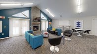 This community room is a well-appointed, multi-functional gathering space designed to accommodate a variety of social and recreational activities. The room is anchored by a striking floor-to-ceiling stacked stone fireplace topped with a wall-mounted television, set beneath soaring vaulted ceilings that create an open, airy atmosphere. Teal accent walls complement the neutral tones throughout, adding a modern, energetic feel to the space.

The lounge area is furnished with vibrant teal sofas and chairs accented with yellow throw pillows, along with sleek black swivel chairs and low-profile coffee tables — creating a comfortable and stylish setting for conversation and relaxation. Natural light filters in through multiple large windows dressed with vertical blinds, brightening the interior throughout the day.

A dedicated recreation zone features a full-size pool table set on patterned carpet that runs throughout the space. Nearby, round café-style tables with black chairs provide casual seating for small groups. A water cooler station adds everyday convenience.

A built-in workstation alcove offers a computer desk and chair, ideal for those needing a quiet spot for work or study. Contemporary linear light fixtures provide consistent, even illumination across all areas. The overall design balances functionality with a welcoming, community-oriented ambiance.