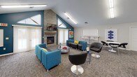 This community room is a well-appointed, multi-functional gathering space designed to accommodate a variety of social and recreational activities. The room is anchored by a striking floor-to-ceiling stacked stone fireplace topped with a wall-mounted television, set beneath soaring vaulted ceilings that create an open, airy atmosphere. Teal accent walls complement the neutral tones throughout, adding a modern, energetic feel to the space.

The lounge area is furnished with vibrant teal sofas and chairs accented with yellow throw pillows, along with sleek black swivel chairs and low-profile coffee tables — creating a comfortable and stylish setting for conversation and relaxation. Natural light filters in through multiple large windows dressed with vertical blinds, brightening the interior throughout the day.

A dedicated recreation zone features a full-size pool table set on patterned carpet that runs throughout the space. Nearby, round café-style tables with black chairs provide casual seating for small groups. A water cooler station adds everyday convenience.

A built-in workstation alcove offers a computer desk and chair, ideal for those needing a quiet spot for work or study. Contemporary linear light fixtures provide consistent, even illumination across all areas. The overall design balances functionality with a welcoming, community-oriented ambiance.