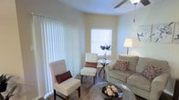 This single-floor residence offers a warm and inviting atmosphere defined by its soft neutral color palette and thoughtfully designed layout. The living area is the centerpiece of the space, bathed in natural light that filters through large vertical blinds and a secondary window fitted with horizontal blinds. The room's bright, creamy walls complement the rich hardwood-style flooring, creating a cohesive and comfortable aesthetic throughout.

The living area is generously proportioned, providing ample room for a full seating arrangement. A plush sofa with decorative accent pillows anchors one side of the room, while a pair of elegant tufted accent chairs add a refined touch. A glass-top coffee table and a petite bistro-style side table with a floral centerpiece offer both function and style. A floor lamp provides warm supplemental lighting, and a ceiling fan ensures year-round comfort.

The large sliding door with vertical blinds suggests access to an outdoor area, enhancing the sense of openness. Overall, the space combines practical design with a light, airy ambiance, making it a welcoming and functional environment suited for everyday living and relaxed entertaining.