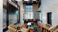 This contemporary clubhouse is defined by its striking architectural design and thoughtfully curated interiors. The centerpiece is a soaring double-height lobby lounge bathed in natural light from expansive floor-to-ceiling gridded windows dressed with striped drapery. Polished concrete floors reflect the ambient glow of sculptural pendant and chandelier lighting, while white painted brick columns and textured wall panels add depth and contrast. The lounge is furnished with bold, patterned armchairs and a dark sectional sofa arranged around a vivid geometric area rug in orange and blue tones, creating an inviting and energetic social atmosphere. Accent seating areas with high-back chairs and rounded side tables are tucked near the textured feature wall, offering quieter spots for relaxation. A dedicated conference room provides a professional setting, featuring a large white resin-topped table, black ergonomic chairs, a wood-planked accent wall with a wall-mounted display, a geometric patterned carpet, and sheer gold drapery. The wet bar area is outfitted with dark granite countertops, stainless steel appliances, and a glamorous gold penny-tile mosaic backsplash lined with display shelving. Additional lounge space with wood flooring and casual seating completes this well-appointed amenity floor.