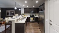This model unit at 7800 Westside in San Diego presents a thoughtfully designed single-floor residence that blends contemporary style with functional living. The open-concept layout flows seamlessly from the kitchen through the dining area and into the living room, all finished with warm wood-look flooring throughout.

The kitchen is a standout feature, outfitted with rich dark espresso cabinetry, bright white countertops, stainless steel appliances including a gas range and refrigerator, and recessed lighting with pendant fixtures illuminating a breakfast bar — ideal for casual dining or entertaining.

The living room is anchored by a bold charcoal accent wall and large sliding glass doors that open onto a private balcony with tropical palm views, filling the space with natural light. The bedroom continues the design language with a striking deep blue accent wall, plush carpeting, a window overlooking lush greenery, and a dedicated walk-through closet with open shelving leading directly to the full bathroom.

The bathroom is generously proportioned, featuring wood-look flooring, dark vanity cabinetry with white countertops, a soaking tub, and ample storage. Throughout the residence, a cohesive palette of navy, charcoal, and warm neutrals creates a sophisticated yet welcoming atmosphere.