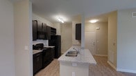 This single-floor unit offers a thoughtfully designed open-concept layout that seamlessly connects the kitchen and living area, creating a spacious and functional environment. The kitchen is a standout feature, showcasing rich dark cabinetry that contrasts elegantly with light granite-style countertops. A peninsula with a built-in stainless steel sink serves as both a workspace and a casual dining or gathering spot, opening directly into the living area. Black appliances, including a gas range, microwave, and refrigerator, complement the kitchen's modern aesthetic. A classic white subway tile backsplash adds a clean, polished touch to the cooking space. Track lighting fixtures illuminate both the kitchen and living areas, ensuring a bright and welcoming atmosphere. Warm-toned, wood-look luxury vinyl flooring runs throughout the unit, providing a cohesive and low-maintenance surface underfoot. Neutral wall tones keep the space feeling open and versatile, ready to accommodate a variety of décor styles. A dedicated entryway near the kitchen offers a practical and well-defined point of entry, contributing to the overall flow of the floor plan. Clean lines and contemporary finishes define the overall character of this well-appointed space.
