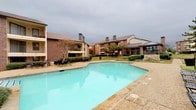 This residential community, identified as Avantic, offers a thoughtfully designed outdoor environment centered around a generously sized swimming pool with a built-in volleyball net, making it a standout amenity for active residents. The pool area is framed by a flagstone and aggregate concrete surround, tiered stone retaining walls with decorative plantings, and multiple lounge chairs for relaxation.

The grounds are well-maintained, featuring manicured lawns, trimmed hedgerows, mature trees, and landscaped garden beds that line the winding walkways connecting the community. The multi-story residential buildings are constructed with a combination of brick and wood siding with pitched rooflines, multiple chimneys, and private balconies offering views of the courtyard and pool area.

A dedicated clubhouse building anchors part of the community, featuring a low-pitched standing-seam metal roof, large multi-pane windows, and an open covered porch with aggregate flooring and outdoor seating. The clubhouse entry provides a bright, welcoming space with views directly into the landscaped courtyard. Exterior utility areas along building facades are neatly integrated with seating nooks and access points, maintaining the community's cohesive and comfortable aesthetic throughout.
