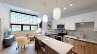 This thoughtfully designed single-floor residence offers a cohesive blend of modern aesthetics and functional living. The open-concept kitchen and living area serves as the heart of the home, where a large central island with a quartz countertop doubles as a casual dining and prep space, illuminated by stylish ribbed globe pendant lights. The kitchen is outfitted with stainless steel appliances, including a French door refrigerator, electric range, microwave, and dishwasher, complemented by a two-tone cabinetry design pairing crisp white uppers with warm wood lowers and a subtle tile backsplash. The living area flows naturally from the kitchen, featuring large windows dressed with roller shades that invite natural light while maintaining privacy. Light hardwood-style flooring runs consistently throughout, tying the spaces together seamlessly. The bedroom is a calm retreat, lined with expansive windows and blackout roller shades for restful sleeping. Generous wall space accommodates a full dresser and mirror. The bathroom features a warm wood vanity with an undermount sink, a well-lit mirror, and a full tub-shower combination, with direct access to the bedroom for added convenience. Clean white walls and recessed lighting throughout the residence contribute to a bright, airy, and contemporary ambiance.