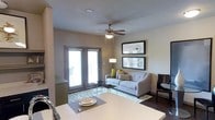 This single-floor space offers a thoughtfully designed open-concept layout that seamlessly connects the living and dining areas. The living room is anchored by a contemporary gray sofa accented with patterned and lime-green throw pillows, set atop a geometric-print area rug that adds visual interest to the dark hardwood flooring. A classic ceiling fan with an integrated light fixture overhead provides both comfort and illumination, while a stylish floor lamp adds warm ambient lighting to the corner of the room. Framed artwork adorns the neutral-toned walls, lending a curated, polished feel to the space. A glass-top coffee table and side table round out the seating area with a sleek, modern touch. Large double doors with blinds open to a private balcony, allowing natural light to filter into the room and offering a connection to the outdoors. The dining area, visible in the foreground, features a clean white surface with place settings, positioned adjacent to the kitchen with a modern faucet detail. The overall ambiance is contemporary and inviting, with a neutral color palette punctuated by carefully chosen accent colors and decor throughout.