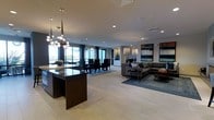 This elegantly designed single-floor residence showcases a seamlessly integrated open-concept layout that unifies the kitchen, dining, and living areas into one expansive, flowing space. Light-toned large-format tile flooring runs throughout, lending a clean, cohesive aesthetic underfoot.

The kitchen anchors one end of the space, featuring a substantial dark-finished island that serves as a striking visual counterpoint to the lighter surroundings. Above, a sculptural multi-globe pendant chandelier adds artistic flair and warm illumination. The dining area sits adjacent, bathed in natural light streaming through floor-to-ceiling windows and glass doors that frame views to the outdoors. Dark dining chairs surround a generously sized table, creating an inviting setting for entertaining.

The living area occupies the opposite end of the open floor plan, centered around a plush sectional sofa in a rich charcoal tone, complemented by a round wood coffee table and a patterned area rug in cool blue tones. Bold abstract artwork adorns the walls, adding a gallery-quality dimension to the space. Recessed lighting throughout ensures consistent, well-distributed illumination, while the abundance of windows creates a bright, airy ambiance that reinforces the home's contemporary and sophisticated character.