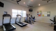 This single-story fitness space is designed to provide a practical and comfortable environment for cardiovascular exercise. The room features a selection of cardio equipment, including treadmills and elliptical trainers, arranged to allow ample space for movement between machines. Neutral-toned carpet covers the floor, contributing to a clean and understated aesthetic throughout the space.

Two ceiling fans are installed overhead, ensuring consistent air circulation during workouts, while recessed lighting provides even illumination across the room. A wall-mounted flat-screen television is positioned for easy viewing from the cardio equipment, enhancing the workout experience. Windows along one wall allow natural light to filter into the space, creating a bright and welcoming atmosphere.

The walls are finished in a soft, neutral tone, kept largely uncluttered to maintain an open feel. A colorful geometric wall art piece adds a touch of visual interest and personality to the space. Overall, the fitness room combines functional equipment with a clean, well-lit design that supports an effective and enjoyable exercise environment.