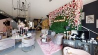 Located at 600 Embarcadero in Carlsbad, this stylish single-floor boutique space blends industrial architecture with glamorous, feminine design. The main retail floor immediately captures attention with its signature floor-to-ceiling floral wall adorned with lush pink and red roses set against a backdrop of green hedging, accented by a neon "#BlushMadeeMeDoIt" sign. Exposed silver ductwork runs overhead, contrasting beautifully with the bold dark charcoal and blush pink color-blocked walls that define the primary lounge and display area. Polished concrete floors flow throughout, lending a clean, modern foundation to the richly layered décor. Seating vignettes are thoughtfully arranged with plush velvet and upholstered chairs, mirrored and glass coffee tables, and an array of curated home goods and lifestyle accessories. A natural wood island counter serves as a central focal point, complemented by open wood shelving lined with décor objects and textiles. A dramatic black staircase with floral garland accents adds vertical interest and leads to additional space above. Pendant and track lighting create a warm, inviting ambiance throughout. A private bathroom features a pedestal sink, grab bar, and decorative wall art, offering a clean and functional amenity. Large storefront windows allow abundant natural light to flood the interior, creating an open, welcoming atmosphere ideal for retail or creative use.