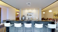 This single-floor space presents a contemporary design centered around a stylish bar or café-style counter finished in dark slate-toned mosaic tile. Three white modern bar stools with chrome frames line the front of the counter, offering casual seating. Behind the counter, a crisp white backsplash and built-in shelving display beverages, cups, and small decorative accents, alongside a coffee machine and sink, suggesting a hospitality or lounge function. Recessed lighting illuminates the central area with a clean, bright ambiance, while pendant lights visible in the adjoining rooms add warm accent lighting. The white walls and ceilings create a sense of openness throughout. To the left, a softly lit room with a low-hung pendant and warm tones hints at a relaxed lounge or seating area. To the right, a glimpse of upholstered furniture suggests an additional sitting space. The overall aesthetic is polished and modern, combining neutral tones with subtle design details to create a welcoming, functional environment suited to social gathering or hospitality use.