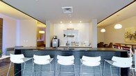 This single-floor space presents a contemporary design centered around a stylish bar or café-style counter finished in dark slate-toned mosaic tile. Three white modern bar stools with chrome frames line the front of the counter, offering casual seating. Behind the counter, a crisp white backsplash and built-in shelving display beverages, cups, and small decorative accents, alongside a coffee machine and sink, suggesting a hospitality or lounge function. Recessed lighting illuminates the central area with a clean, bright ambiance, while pendant lights visible in the adjoining rooms add warm accent lighting. The white walls and ceilings create a sense of openness throughout. To the left, a softly lit room with a low-hung pendant and warm tones hints at a relaxed lounge or seating area. To the right, a glimpse of upholstered furniture suggests an additional sitting space. The overall aesthetic is polished and modern, combining neutral tones with subtle design details to create a welcoming, functional environment suited to social gathering or hospitality use.