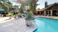 Located at 8017 Ashberry Lane in Pasadena, Maryland, this single-level outdoor amenity space is centered around a generously sized swimming pool with clear blue water and tiled pool edge detailing. The pool is flanked by a broad, stone-paved sundeck that provides ample room for relaxation and recreation. Surrounding the pool area is an extensive collection of lounge chairs, including cushioned chaise lounges in vibrant teal, as well as additional seating arrangements with side tables for comfort and convenience. A rustic wooden pergola anchors one end of the deck, offering a shaded retreat for those seeking relief from the sun. The landscape design is a standout feature, with mature trees — including flowering crape myrtles and palms — providing natural shade and visual appeal throughout the space. Large decorative planters filled with blooming flowers add elegant touches along the poolside. A wrought iron fence encloses the area, ensuring privacy and security while maintaining an open, airy feel. Adjacent to the pool, a clubhouse-style structure with a pitched roof, large windows, and a covered patio provides additional indoor-outdoor gathering space. The overall ambiance is resort-like, combining well-maintained greenery, quality hardscaping, and thoughtful amenities to create an inviting and functional outdoor environment.