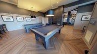 This single-floor clubhouse is thoughtfully designed to offer a variety of social and functional spaces, each finished with a cohesive modern aesthetic. Herringbone-patterned hardwood flooring runs throughout, tying together every area with warmth and visual interest.

The billiards area serves as a central gathering point, housing two full-sized pool tables beneath large cylindrical pendant lights. Adjacent to it, a sophisticated lounge features bold blue velvet seating arranged around a dramatic floor-to-ceiling dark tile fireplace, creating an inviting focal point. Recessed lighting and elevated ceilings amplify the sense of openness throughout.

A quieter sitting area with white wainscoting, grey upper walls, and a tray ceiling offers a more intimate retreat, furnished with upholstered swivel chairs and accented with framed artwork.

The clubhouse kitchen is equipped with grey cabinetry, dark countertops, stainless steel appliances, and a striking vertical moss wall accent alongside white shiplap tile, adding a natural, organic element to the contemporary design.

A private conference room with large windows, floor-length drapes, and a dark meeting table provides a dedicated workspace. Natural light filters through generously sized windows, enhancing the overall welcoming ambiance of the clubhouse.