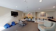 This single-floor communal space is thoughtfully designed to accommodate a variety of activities, from relaxed socializing to focused work. The main lounge area features warm, neutral-toned walls accented by colorful abstract artwork, creating an inviting yet functional atmosphere. Recessed lighting throughout ensures consistent illumination across the space.

The layout is divided into distinct zones. A comfortable L-shaped sectional sofa anchors a lounge seating area, accompanied by decorative accent tables and tall indoor plants that add a touch of greenery. A wall-mounted flat-screen TV serves this relaxed gathering area. Adjacent to it, a round table with chairs provides casual meeting space, while a row of yellow counter-height stools lines a built-in wall-mounted desk, ideal for individual work or study.

A secondary counter-height workstation with white metal stools occupies a separate nook, distinguished by light tile flooring that transitions from the patterned carpet found throughout the main area.

The kitchenette is equipped with dark cabinetry, granite-style countertops, a double stainless steel sink, a full-size refrigerator, and a microwave, supporting everyday convenience. Natural light enters through a window near the lounge seating, contributing to the overall welcoming ambiance of the space.