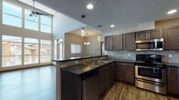 Located at 17016 East Indiana Parkway in Spokane Valley, WA, this single-floor unit offers a thoughtfully designed open-concept layout highlighted by dramatically vaulted ceilings that create an impressive sense of volume and airiness throughout. The living area is bathed in abundant natural light streaming through a towering wall of floor-to-ceiling windows, complemented by a ceiling fan overhead and rich, dark hardwood-style flooring that runs throughout the main living spaces. A private exterior door provides direct access from the living area, further enhancing the open feel.

The kitchen is well-appointed with dark rich cabinetry, dark countertops, a stone tile backsplash, and a full suite of stainless steel appliances including an electric range, over-the-range microwave, and dishwasher. Pendant lighting above the kitchen peninsula adds a modern touch, while the open pass-through connects seamlessly to the living and dining areas.

The bedroom features plush carpeting and its own vaulted ceiling section, with a window offering views to the outside. The bathroom includes a soaking tub and wood-style flooring. A dedicated laundry room houses a full-size washer and dryer with overhead shelving for added storage convenience.
