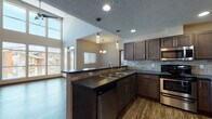Located at 17016 East Indiana Parkway in Spokane Valley, WA, this single-floor unit offers a thoughtfully designed open-concept layout highlighted by dramatically vaulted ceilings that create an impressive sense of volume and airiness throughout. The living area is bathed in abundant natural light streaming through a towering wall of floor-to-ceiling windows, complemented by a ceiling fan overhead and rich, dark hardwood-style flooring that runs throughout the main living spaces. A private exterior door provides direct access from the living area, further enhancing the open feel.

The kitchen is well-appointed with dark rich cabinetry, dark countertops, a stone tile backsplash, and a full suite of stainless steel appliances including an electric range, over-the-range microwave, and dishwasher. Pendant lighting above the kitchen peninsula adds a modern touch, while the open pass-through connects seamlessly to the living and dining areas.

The bedroom features plush carpeting and its own vaulted ceiling section, with a window offering views to the outside. The bathroom includes a soaking tub and wood-style flooring. A dedicated laundry room houses a full-size washer and dryer with overhead shelving for added storage convenience.