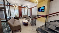Located at 4401 East-West Hwy in Bethesda, Maryland, this single-floor commercial lobby presents a polished and professional environment defined by high-end architectural finishes and thoughtful design. The main entrance features striking polished marble flooring in contrasting cream and deep green tones, complemented by gold-accented ceiling details and a series of distinctive starburst chandeliers that lend a contemporary yet classic character to the space.

A dedicated reception desk anchors the central lobby area, which also features an intricately patterned carpet with swirling organic motifs in navy and gold tones. Rich mahogany millwork and column detailing run throughout, providing warmth and visual continuity. Gold elevator doors and wall panel accents further reinforce the upscale aesthetic.

A comfortable lounge area is thoughtfully appointed with plush sofas, patterned accent chairs, and a round cocktail table, set beneath a sputnik-style chandelier. Floor-to-ceiling windows in this area allow abundant natural light and outdoor views. A wall-mounted flat-screen television and a decorative fireplace accent wall add functional and visual interest to the lounge.

Additional corridors lead to mailbox banks and secondary stairwells, finished consistently with the same quality materials. Overall, the space conveys a refined, welcoming professional atmosphere suited for a premier Bethesda commercial address.