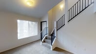 This two-story space is defined by clean, neutral interiors and a straightforward layout connecting both floors. The main level features wall-to-wall carpeting in a neutral tone, contributing to a comfortable and cohesive feel throughout. A large window fitted with horizontal blinds allows natural light to filter into the living area, brightening the white walls and creating an open, airy atmosphere.

A centrally positioned staircase serves as a functional focal point, featuring carpeted treads that match the flooring, white-painted wood bases, and striking black metal balusters paired with light wood handrails. The staircase design is modern yet understated, complementing the overall minimalist aesthetic of the space. The upper level is visible from below, with matching black metal railings lining the upper landing, maintaining design consistency across both floors.

The entry area includes a white paneled door, providing a clean and welcoming threshold into the home. Ceiling-mounted light fixtures on both levels provide additional illumination. The white walls throughout create a bright, neutral backdrop well-suited for a variety of interior styles.
