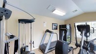 This single-floor fitness room is designed to provide a functional and efficient workout environment. The space is outfitted with a thoughtful selection of exercise equipment, including a treadmill, an elliptical machine, and a multi-function cable weight machine, allowing for a comprehensive range of cardiovascular and strength training exercises. A flat-screen display is mounted nearby, offering entertainment or instructional viewing during workouts. The room features neutral, light-colored walls that contribute to an open and clean atmosphere, while flush-mounted fluorescent ceiling lights ensure the space is well-lit throughout. Carpeted flooring adds a layer of comfort underfoot. A framed motivational print on the wall adds a finishing decorative touch. A window allows for some natural light to filter into the room, enhancing the overall ambiance. The layout is organized and practical, making efficient use of the available space to accommodate multiple users and exercise modalities. Overall, this fitness room serves as a convenient, self-contained facility suited for daily exercise needs.