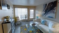 This single-floor living space showcases a well-appointed living room designed with a clean, contemporary aesthetic. The room features hardwood-style flooring in a warm grey-brown tone, providing a sleek and durable foundation throughout. Large windows dressed with sheer white curtains allow generous natural light to fill the space, complemented by a glass-panel entry door that further brightens the interior.

The living area is anchored by a plush white leather sofa adorned with decorative pillows in teal and neutral tones, paired with a transparent glass coffee table that enhances the open, airy feel of the room. A patterned area rug in grey and yellow adds a bold pop of color and defines the seating zone. A modern accent chair with a chrome frame and white cushioning provides additional seating near the window.

Storage and display are addressed by a mirrored accent cabinet along one wall, while a table lamp in teal and a large abstract canvas artwork in blue and grey tones introduce layered visual interest. Subtle decorative touches, including a floral arrangement and a sculptural centerpiece on the coffee table, complete the polished, inviting ambiance of the space.