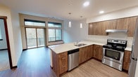 Located at 21044 Viking Way NW in Poulsbo, Washington, this single-floor residence offers a thoughtfully designed layout with contemporary finishes throughout. The kitchen serves as a central highlight, showcasing warm wood-tone cabinetry paired with crisp white countertops and a peninsula that provides additional workspace and casual seating. Stainless steel appliances, including a range, dishwasher, and refrigerator, complement the modern aesthetic, while pendant lighting above the peninsula adds a refined touch. Recessed lighting and wood-look flooring extend throughout the main living and kitchen areas, creating a cohesive and inviting atmosphere.

Large windows in the living area frame views of the surrounding wooded landscape and provide generous natural light. A sliding glass door opens to a private balcony, further connecting the interior to the natural setting outside. The bedrooms feature plush carpeting and ample closet space, with sliding closet doors offering practical storage. One bedroom benefits from an oversized horizontal window that draws in treetop views and abundant daylight, enhanced by a baseboard heating unit below. Neutral wall tones throughout the space provide a clean, versatile backdrop that complements the warm wood and white design elements.