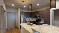 This single-floor residence in Euless, Texas, showcases a thoughtfully designed kitchen as its centerpiece. The kitchen features rich, dark wood cabinetry with a horizontal grain pattern, providing ample storage while contributing to a sleek, modern aesthetic. Light granite countertops with subtle veining offer a striking contrast against the darker cabinetry, and the oversized kitchen island with an undermount stainless steel sink serves as both a functional workspace and a natural gathering point.

Stainless steel appliances, including a French door refrigerator, electric range, and built-in microwave, complement the contemporary design. A classic subway tile backsplash adds texture and visual interest to the cooking area. Recessed lighting is supplemented by two cylindrical pendant lights suspended above the island, providing focused task lighting with a decorative flair.

The kitchen opens to the surrounding living space, promoting an open, connected feel. Warm-toned hardwood-style flooring flows throughout, enhancing the overall cohesion of the space. The combination of modern fixtures, quality materials, and carefully considered lighting gives this residence a polished, move-in-ready appeal.