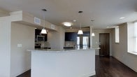 This single-floor space showcases a thoughtfully designed open-concept layout that seamlessly integrates the kitchen and living areas. The kitchen is a standout feature, characterized by dark espresso cabinetry that contrasts beautifully with the light granite countertops. A curved kitchen island serves as the centerpiece, providing generous workspace and a natural gathering point within the space.

Three pendant lights hang above the island, offering focused task lighting while contributing to the modern aesthetic. Stainless steel appliances, including a refrigerator, microwave, and range, are integrated within the cabinetry, reinforcing the sleek, contemporary design. A neutral tile backsplash adds texture behind the cooking area.

Dark hardwood flooring extends throughout the open living space, grounding the design with warmth and continuity. Flush ceiling lighting, combined with the pendant fixtures, ensures the space is well-illuminated. A full-height entry door with modern hardware is visible, adding to the refined character of the unit. Overall, the space delivers a clean, modern ambiance with an emphasis on functional design and quality finishes.